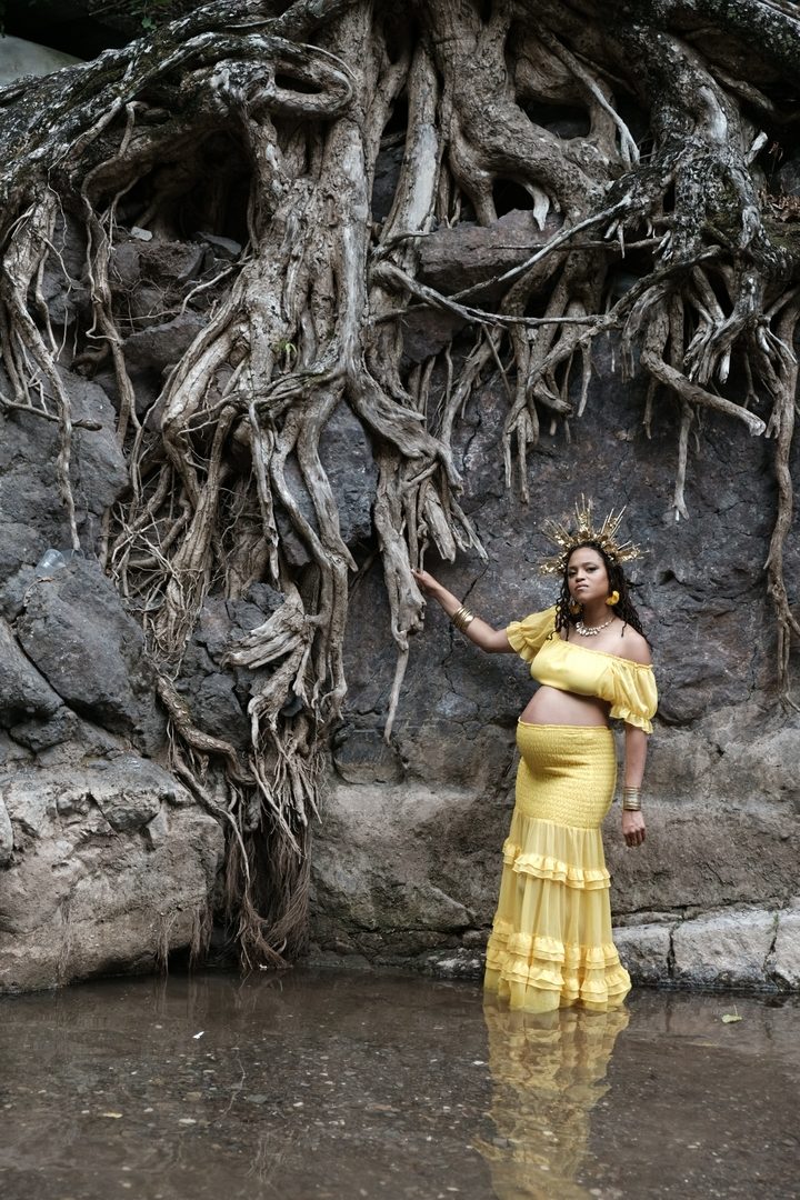 The Charged Rootz stretch therapist in a golden gown and sun crown, standing in shallow water and reaching up into the roots of a great tree